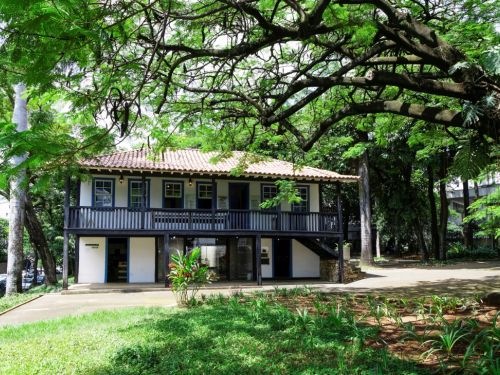 Casa principal do Museu Histórico Abílio Barreto, rodeada por uma área verde. A casa histórica, de dois andares, tem paredes brancas e uma varanda escura no andar de cima. Uma grande árvore com folhagem verde e densa faz uma moldura na parte superior. O jardim tem gramado e um caminho de terra que leva até a casa.