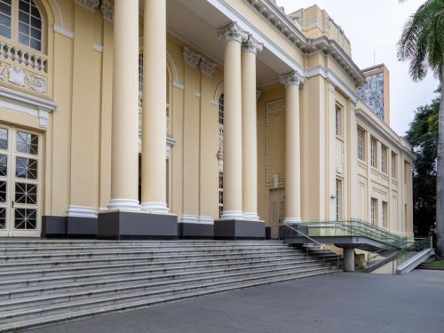 A fachada do BEFLY Minas Centro, em BH, é um edifício clássico creme com grandes colunas brancas e uma ampla escadaria de granito. À direita, uma rampa moderna de vidro e metal oferece acesso. Palmeiras altas e vegetação emolduram a estrutura, com um prédio ao fundo.