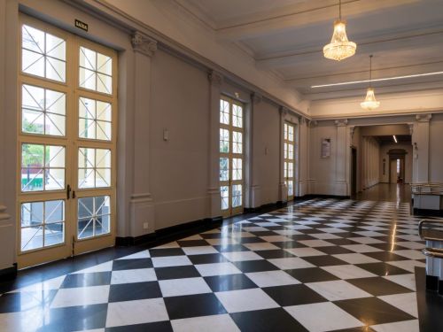 Interior do BEFLY Minas Centro, BH. O chão brilhante exibe um padrão xadrez preto e branco. Grandes portas de vidro na parede esquerda, com "SAÍDA", iluminam o hall. Paredes claras e colunas decoradas conduzem a um corredor elegante ao fundo, sob lustres de cristal e luzes modernas.