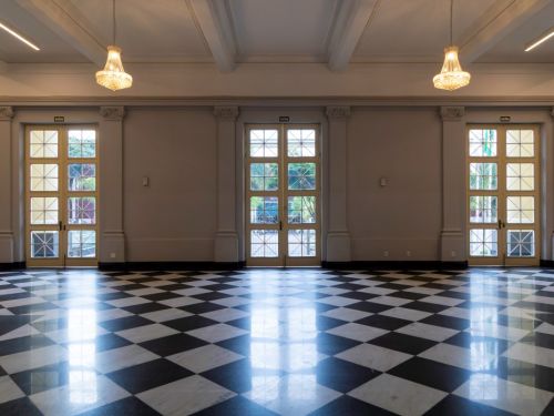 Hall elegante no BEFLY Minas Centro, BH, com chão de xadrez preto e branco brilhante. Três portas duplas de vidro, marcadas "SAÍDA", iluminam o espaço. Lustres de cristal e luzes modernas pendem do teto alto, com paredes claras e colunas decoradas.