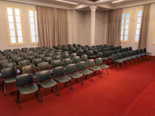 Auditório no BEFLY Minas Centro, BH. Chão de carpete vermelho vivo. Dispostas em fileiras, cadeiras escuras ocupam a sala. Nas paredes claras, janelas altas com molduras claras têm cortinas bege. Uma coluna decorada no canto e iluminação moderna linear no teto completam o ambiente.