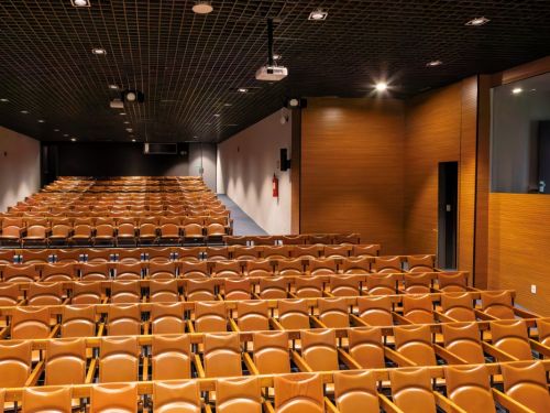 Sala de auditório ampla e vazia no BeFly Minascentro, Belo Horizonte. Vista ampla das fileiras de cadeiras acolchoadas em couro marrom, dispostas em degraus ascendentes, com braços de madeira. Teto preto com grade metálica e luzes embutidas redondas. Projetor branco pendurado no centro. Paredes laterais: esquerda cinza clara com extintor vermelho; direita em painéis de madeira marrom com janela espelhada e porta preta. Iluminação suave, piso azul escuro na frente. Ambiente moderno e acolhedor para eventos.