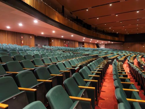 Grande auditório vazio no BeFly Minascentro, Belo Horizonte. Fileiras curvas de cadeiras verdes escuras com braços de madeira, dispostas sobre carpete vermelho. Teto alto com luzes embutidas e painéis de madeira nas paredes e varanda superior. Ambiente silencioso, com extintores vermelhos nas laterais, ideal para grandes eventos ou apresentações teatrais.