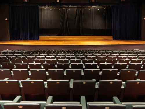 Auditório vazio no BeFly Minascentro, Belo Horizonte. Palco de madeira com cortinas pretas escuras e iluminação suave. Fileiras de cadeiras verde-escuras com braços de madeira, dispostas em carpete vermelho. Teto com luzes embutidas e trilhos. Paredes escuras com detalhes discretos. Ambiente calmo, pronto para apresentações teatrais ou eventos culturais.
