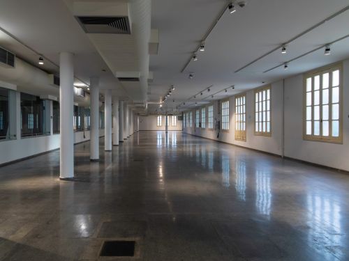 Espaço amplo e vazio no BeFly Minascentro, Belo Horizonte. Piso de pedra polida e escura, refletindo a luz. Colunas brancas alinhadas, teto com dutos de ventilação e luzes embutidas. Paredes brancas com janelas altas de madeira e vidro, permitindo luz natural. Extintores vermelhos nas laterais. Ambiente limpo, ideal para exposições ou eventos.