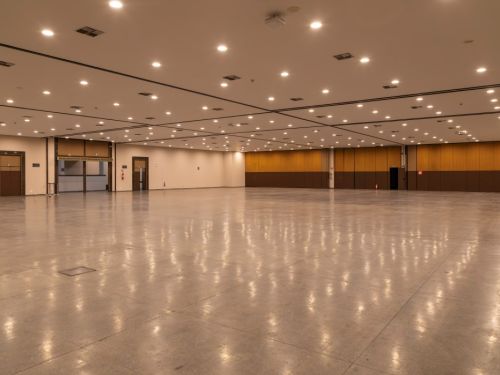 Grande salão vazio no BeFly Minascentro, Belo Horizonte. Piso polido e reflexivo, iluminado por luzes embutidas no teto branco. Paredes com painéis de madeira marrom e portas escuras nas laterais, além de extintores vermelhos. Ambiente amplo e limpo, ideal para feiras, exposições ou eventos corporativos.