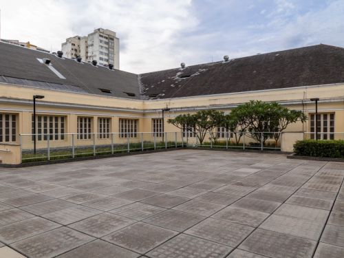 Terraço aberto no BeFly Minascentro, Belo Horizonte. Piso de lajotas cinza em quadrados, cercado por grade metálica. Edifício com paredes amarelo-claro e janelas altas de madeira. Árvores pequenas e arbustos verdes ao fundo, com prédios altos ao longe sob céu nublado. Teto do prédio com telhas escuras e ventiladores. Ambiente calmo e espaçoso.