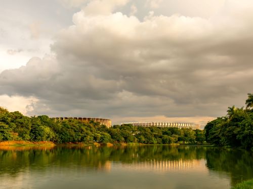 Uma vista ampla do estádio ao fundo, parcialmente encoberto pela vegetação densa e pelo relevo natural. Em primeiro plano, um lago reflete a copa das árvores e o céu dramático, coberto por nuvens pesadas em tons de cinza e dourado, iluminadas pela luz suave do entardecer.