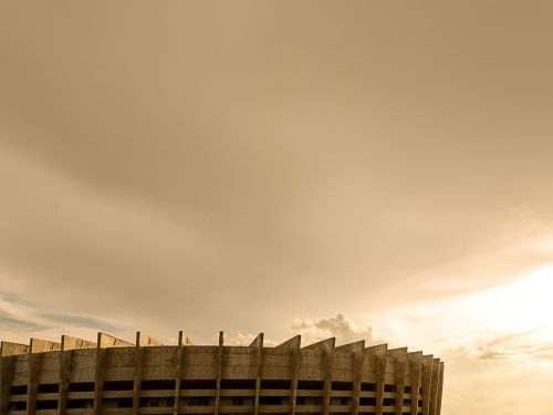 A imagem mostra o topo do estádio Mineirão, em Belo Horizonte, Minas Gerais. A estrutura de concreto, que é uma característica marcante do estádio, é vista em um ângulo de baixo para cima, com um grande céu claro e nuvens esparsas ao fundo. 