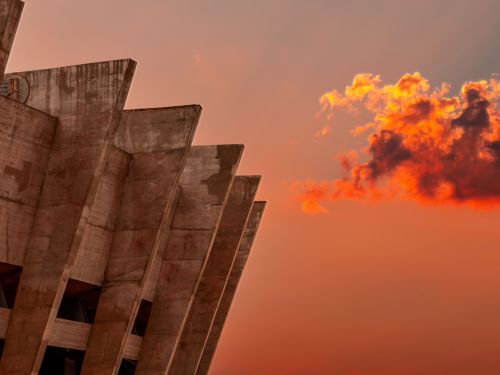 A imagem exibe uma vista lateral de uma seção da arquitetura do estádio Mineirão. Pilares de concreto em formato triangular se projetam do corpo principal do edifício, criando um padrão de sombras e linhas. A cena é banhada pela luz quente do pôr do sol, que tinge o céu de tons alaranjados e avermelhados. À direita, uma grande nuvem escura é iluminada por baixo, parecendo incandescente e contrastando com a estrutura de concreto. 