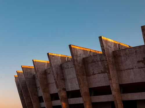 A imagem mostra uma fileira de pilares de concreto, característicos do estádio Mineirão, em um ângulo de baixo para cima. A luz do sol atinge a borda superior dos pilares, criando um brilho dourado que contrasta com o tom cinza e texturizado do concreto. O céu ao fundo está limpo, com um degradê de azul claro que se torna um tom alaranjado próximo ao horizonte, sugerindo o nascer ou pôr do sol. 