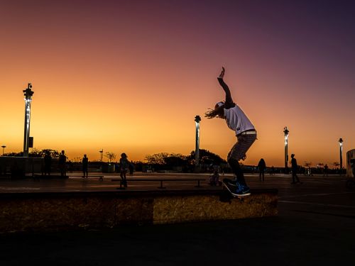 A imagem retrata a silhueta de um skatista em movimento, realizando uma manobra em uma plataforma. Ele está no ar, com os braços erguidos para se equilibrar. O cenário, que está em segundo plano, é uma esplanada iluminada por postes de luz e pessoas praticando o esporte, todos em silhueta contra um céu de pôr do sol com um forte gradiente de cor, indo do amarelo e laranja vibrantes na parte inferior até um roxo e azul escuro no topo.