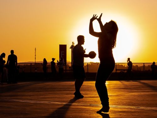 A imagem mostra duas pessoas em silhueta, jogando vôlei ao ar livre durante o pôr do sol. Uma mulher, em primeiro plano, se prepara para receber a bola, que está no alto, enquanto um homem fica em segundo plano. O sol, que está posicionado atrás deles, cria um halo de luz intensa e alaranjada, que se reflete na quadra. Ao fundo, outras pessoas em silhueta e um horizonte com prédios podem ser vistos.