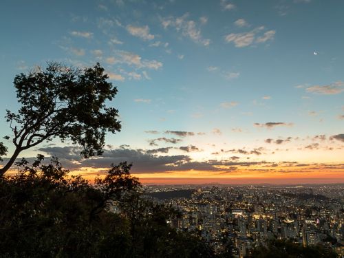 Vista panorâmica da cidade de Belo Horizonte ao entardecer. No primeiro plano, há a silhueta escura de uma árvore e vegetação. Acima, o céu tem tons de laranja e vermelho, que se mesclam com o azul. Uma lua crescente é visível no canto superior direito. Abaixo, a vasta área urbana começa a acender suas luzes, criando um tapete de pontos brilhantes.