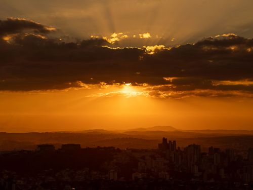 Uma vista do pôr do sol sobre a cidade. O céu está em tons de laranja e vermelho. Raios de sol dourados atravessam nuvens escuras, criando um brilho intenso. Abaixo, a cidade e as montanhas ao fundo estão em silhueta, formando camadas escuras que se estendem até o horizonte.
