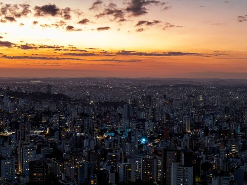 Uma vista panorâmica da cidade ao anoitecer. O céu, no alto, tem tons de laranja e amarelo, com nuvens escuras e iluminadas por baixo. Abaixo, a vasta área urbana se estende até o horizonte, com incontáveis edifícios. As luzes dos prédios começam a brilhar, criando um tapete de pontos luminosos que cobrem toda a paisagem.