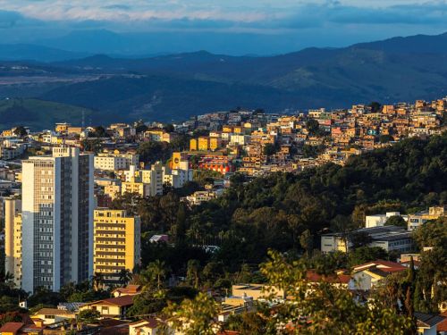 Vista em camadas de uma encosta da cidade. No primeiro plano, um prédio branco e moderno e os telhados de casas. No meio, uma área de morros densamente ocupada por casas de diversas cores, como amarelo e vermelho. Ao fundo, uma cadeia de montanhas escuras se estende no horizonte, sob um céu claro e nebuloso.