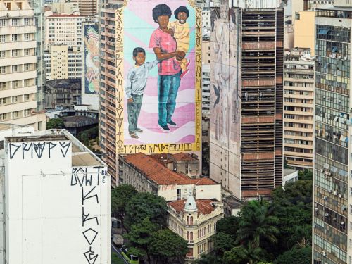 Vista aérea do centro de Belo Horizonte, com edifícios altos e ao fundo a Serra do Curral. Em destaque, um grande mural em fachada de prédio retrata uma pessoa negra segurando uma criança no colo, acompanhada de outra criança ao lado.