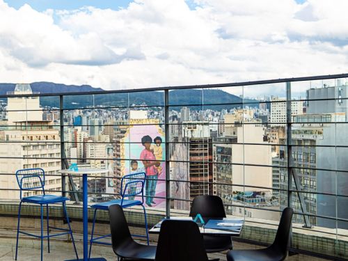 Área externa com mesas e cadeiras pretas e azuis sobre piso de concreto. A cena mostra a vista da cidade com edifícios ao fundo e, em destaque, o mesmo mural de família negra em fachada de prédio, visível através do guarda-corpo envidraçado.