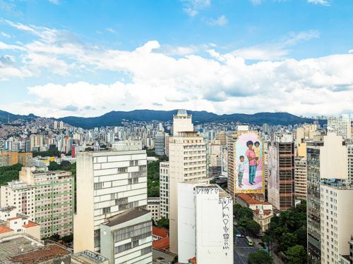 Panorâmica da cidade de Belo Horizonte, com vários prédios de diferentes alturas. Em destaque, o mural colorido em fachada retratando uma pessoa negra segurando uma criança, com outra ao lado. Ao fundo, a Serra do Curral e céu parcialmente nublado.
