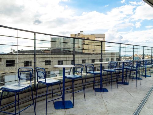 Espaço externo com mesas e cadeiras azuis alinhadas junto ao guarda-corpo de vidro. Ao fundo, prédios de fachada clara e janelas pequenas, em primeiro plano um edifício revestido de pastilhas. Céu com nuvens claras ao fundo.