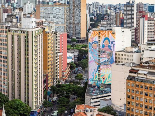 Vista de avenida central de Belo Horizonte com tráfego leve, prédios altos e edifício com mural colorido mostrando duas figuras humanas. Ao redor, fachadas residenciais e comerciais em tons variados. Céu parcialmente encoberto por nuvens claras.
