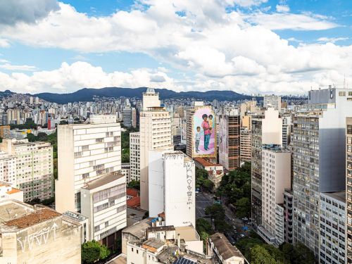 Panorâmica da cidade com destaque para edifício em cuja fachada está o mural colorido de pessoa negra segurando criança, acompanhado de outra criança. Serra do Curral aparece ao fundo, sob céu azul com nuvens brancas.