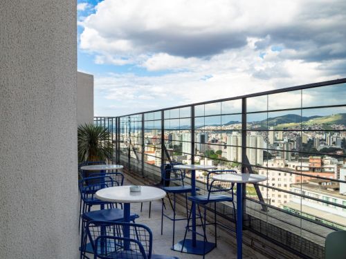 Sacada externa com mesas redondas brancas e cadeiras azuis metálicas sobre piso de concreto. Proteção lateral de vidro transparente, mostrando ao fundo edifícios altos de Belo Horizonte sob céu parcialmente nublado.