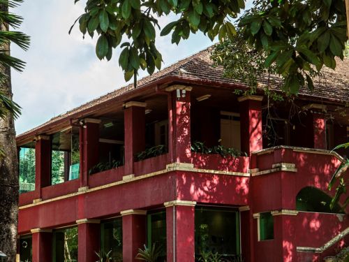 Prédio de dois andares, pintado de vermelho escuro com detalhes em bege claro. Possui varandas com colunas e floreiras, e um telhado de telhas cerâmicas. Está rodeado por árvores grandes e vegetação, com uma calçada de pedras levando à entrada.