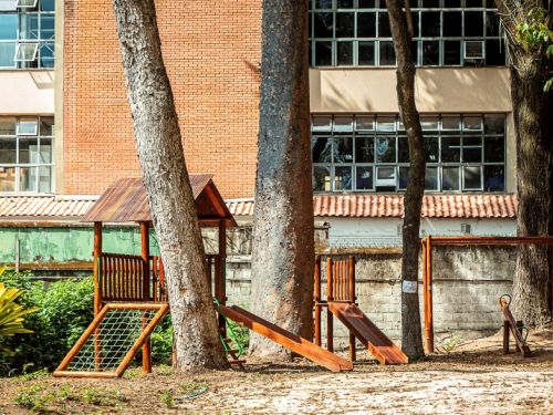 Dito pelo ChatGPT: A imagem mostra um parquinho infantil em área externa sombreada por árvores altas. Os brinquedos, feitos de madeira, incluem escorregador, balanço, escada de cordas e gangorra, todos posicionados sobre chão de terra batida. Ao fundo, observa-se um edifício de tijolos aparentes e grandes janelas envidraçadas.