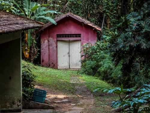 Dito pelo ChatGPT: A imagem mostra uma casa simples, pintada de vermelho, com portas de madeira brancas, localizada em uma trilha no meio de uma vegetação densa e verde. Ao redor, árvores e plantas tropicais formam um cenário de floresta. O caminho de pedras leva até a estrutura.