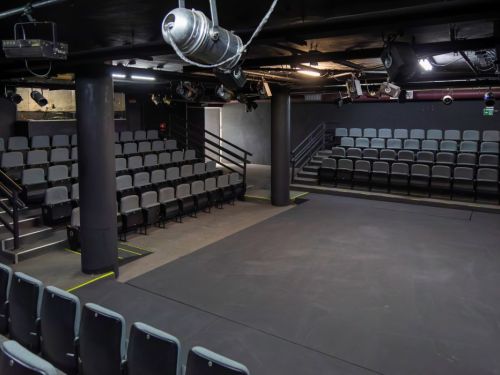 Interior de um teatro pequeno, com todas as paredes, teto e chão pintados de preto. O local tem duas arquibancadas com fileiras de cadeiras cinzas, com corrimões de metal. O palco, no centro, é uma área plana e escura. O teto tem tubulações e equipamentos de iluminação visíveis.