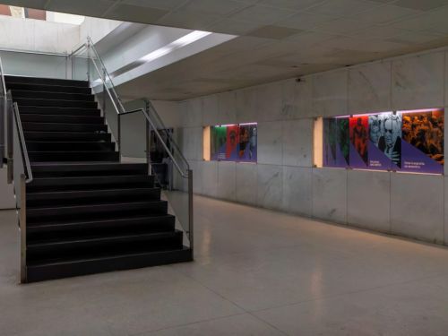 O interior do Palácio das Artes, com uma escadaria de degraus escuros e corrimão de metal e vidro à esquerda. À direita, um longo corredor com paredes de mármore branco. Na parede, uma série de painéis retangulares iluminados por dentro exibe fotos coloridas de pessoas. O ambiente é bem iluminado por luzes no teto.