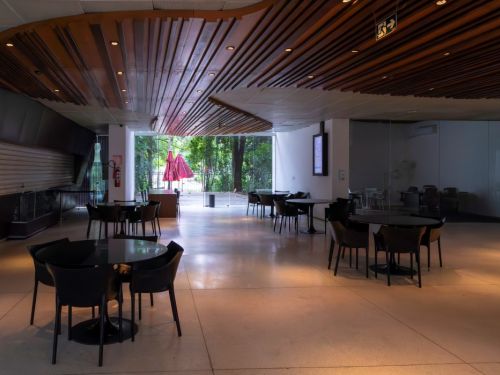 O interior de um café ou lounge vazio. O espaço tem várias mesas redondas e cadeiras pretas. O teto tem um design moderno, com ripas de madeira e luzes embutidas. No fundo, uma grande parede de vidro dá vista para uma área verde com árvores. Há um guarda-sol vermelho do lado de fora. O ambiente tem uma iluminação suave e quente.