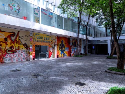 O pátio externo do Palácio das Artes. O chão é de pedras claras. A parede do prédio, no fundo, é coberta por um grande e colorido mural de grafite, com figuras e formas abstratas. Acima do mural, a parte de cima do prédio tem janelas de vidro. Árvores com troncos escuros e copas verdes estão espalhadas pelo local, fazendo sombras no chão.