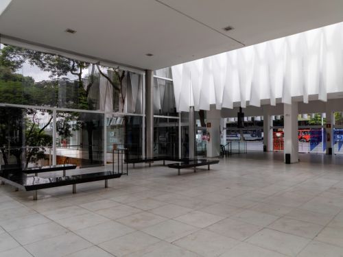 O interior de um grande salão moderno e vazio. O chão é de piso claro. No centro, há bancos pretos e longos. O teto tem uma estrutura branca de formas verticais. Uma parede de vidro à esquerda oferece vista para árvores. À direita, uma grande abertura dá para a rua, onde é possível ver veículos e um ônibus vermelho.