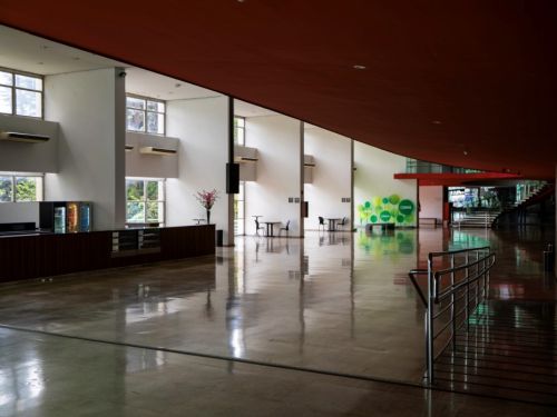 Um grande e moderno salão. O chão é um piso claro e muito brilhante, que reflete as luzes e o ambiente. O teto é alto, curvo e inclinado, na cor vermelho terracota. À esquerda, há um balcão com janelas que dão para a área externa. No fundo, uma escada em espiral e, na parede, um logo colorido.