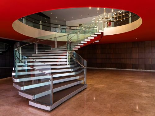 Uma escadaria moderna em formato de espiral. Os degraus brancos, com corrimão de vidro e metal, sobem do chão de pedra clara para um andar superior. O teto, na cor vermelha, tem uma grande abertura circular. A parede do fundo é revestida de madeira escura, fazendo um contraste com o ambiente claro e moderno.