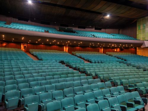 Imensa plateia vazia do Grande Teatro Palácio das Artes. O espaço é preenchido por inúmeras fileiras de poltronas em um tom de azul-esverdeado, dispostas em vários blocos com corredores. Há um balcão com mais poltronas no andar de cima. As paredes são de madeira escura e o teto é preto e curvo, com luzes embutidas que iluminam o local.