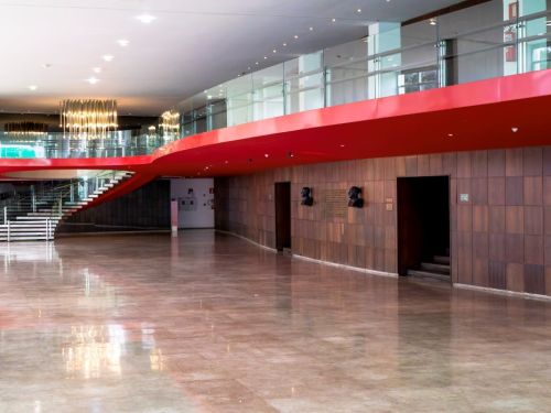 O saguão principal do Palácio das Artes. O chão é de mármore claro e brilhante. À esquerda, uma larga escadaria leva a um andar superior. A parede do fundo é coberta por painéis de madeira marrom, com duas esculturas em formato de busto. O teto e a varanda interna são vermelhos, fazendo um forte contraste no ambiente.