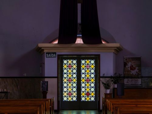 O interior de uma igreja, com foco no fundo do corredor. No centro, uma porta dupla de vitral com círculos coloridos em amarelo, azul e vermelho, por onde a luz entra. O chão de pedra escura e brilhante reflete a luz. De cada lado, há fileiras de bancos de madeira. Acima da porta, um crucifixo está coberto por um tecido roxo.