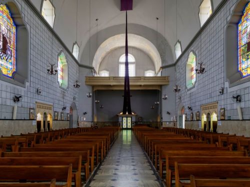 O interior de uma igreja. A vista é de um longo corredor central com chão de azulejos claros e escuros, ladeado por fileiras de bancos de madeira. As paredes têm janelas grandes de vitral coloridos e o teto é alto e em formato de abóbada. No fundo, há uma varanda de madeira e um tecido roxo pendurado, que descem em direção ao chão.