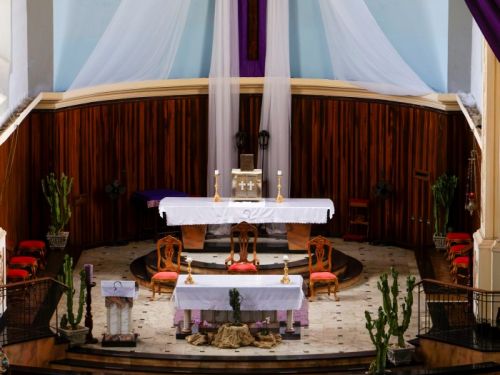 Altar de uma igreja católica, em uma área elevada com vários degraus. Grandes tecidos roxos e brancos pendem do teto, emoldurando uma cruz de madeira. A parede do fundo é de madeira escura. Há dois altares cobertos por tecidos brancos, com cadeiras e vasos de cactos ao lado. No primeiro plano, estão as primeiras fileiras dos bancos da igreja.