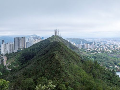 Vista panorâmica da cidade de Belo Horizonte. A serra do curral coberta por folhagem verde e densa divide a paisagem. De um lado, há um grupo de prédios altos e brancos. Do outro, a vasta paisagem urbana se estende até o horizonte. No topo da montanha central, há várias antenas. O céu está coberto por nuvens brancas e cinzas.