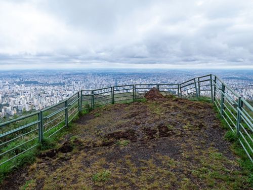 Vista panorâmica da cidade de Belo Horizonte, tirada de um mirante no Parque Serra do Curral. O local é cercado por uma grade de metal verde. A terra do mirante é escura com grama. Ao fundo, a vasta paisagem da cidade, com inúmeros edifícios, se estende até o horizonte. O céu está totalmente coberto por nuvens cinzas e brancas.