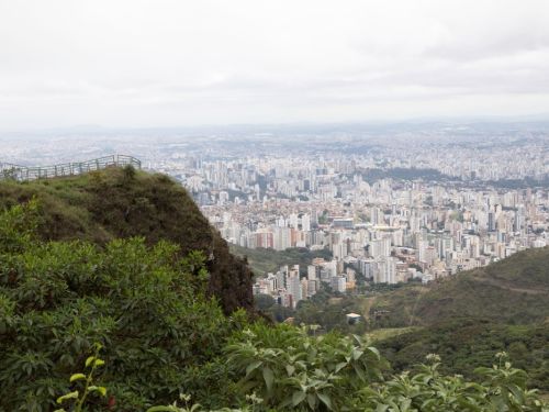 A vasta paisagem da cidade de Belo Horizonte, vista do alto do Parque Serra do Curral. Em primeiro plano, uma encosta íngreme com folhagem verde escura e terra avermelhada. Do outro lado da encosta, a vasta paisagem urbana se estende até o horizonte, com incontáveis edifícios. O céu está totalmente coberto por nuvens cinzas e brancas.