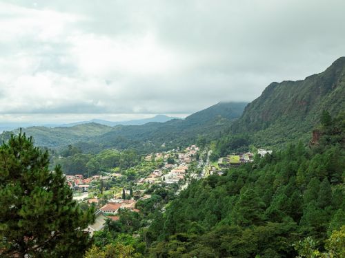 Vista do Parque Serra do Curral, onde um pequeno bairro com casas de telhado vermelho e branco está aninhado. O vale é cercado por montanhas de encostas íngremes e cobertas de folhagem densa. A paisagem é dominada pelo verde e pelo cinza das nuvens, que cobrem o topo das montanhas, tornando a cena suave e nebulosa.