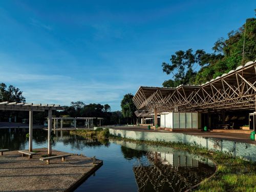 Estrutura arquitetônica com cobertura metálica em frente a um lago. À esquerda, pergolado de concreto com bancos de madeira sobre piso de pedras. Árvores grandes ao fundo, céu azul sem nuvens.