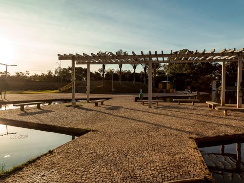 Área aberta com piso de pedras, pergolado de concreto com bancos de madeira e lago refletindo a luz do sol. Ao fundo, árvores e palmeiras alinhadas, com céu claro e ensolarado.