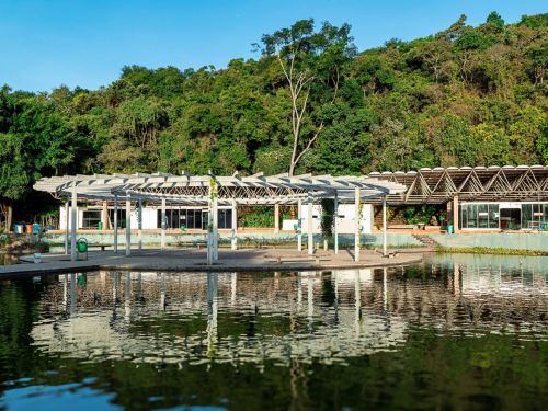 Lago em primeiro plano refletindo estruturas brancas de pergolados e um prédio com cobertura metálica. Atrás do prédio há densa vegetação verde cobrindo uma encosta.